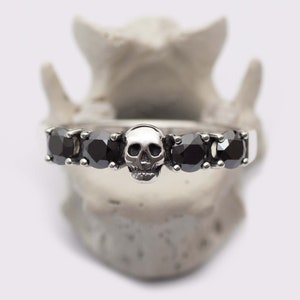 May include: A silver band ring with five black gemstones and a skull design in the center.