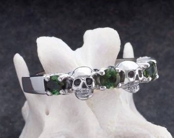 May include: A silver ring with five skull-shaped accents and green gemstones. The ring is on a white bone-like surface.