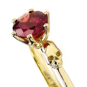 May include: A gold ring with a red gemstone and a skull design. The ring is set with a round, red gemstone in a prong setting. The skull is on the band of the ring and is facing forward.