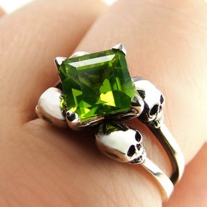 Könnte beinhalten: Ein silberner Ring mit einem grünen Edelstein und zwei Schädeldetails. Der Ring wird an einem Finger getragen.