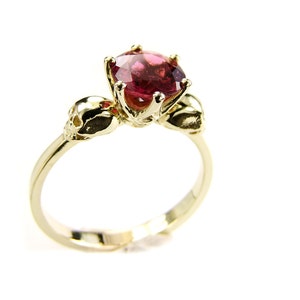 May include: A gold ring with a pink gemstone and two skull accents.