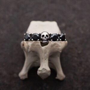 May include: A silver ring with a skull design and black gemstones. The ring is displayed on a white surface.