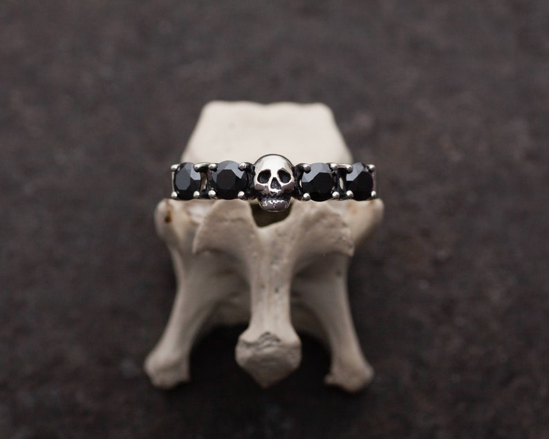 May include: A silver ring with a skull design and black gemstones. The ring is displayed on a white surface.
