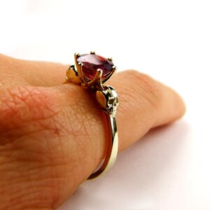 May include: A gold ring with a large red gemstone and two skull accents on the band.