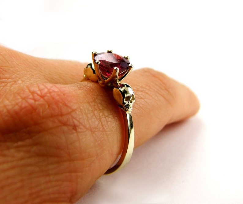 May include: A gold ring with a large red gemstone and two skull accents on the band.