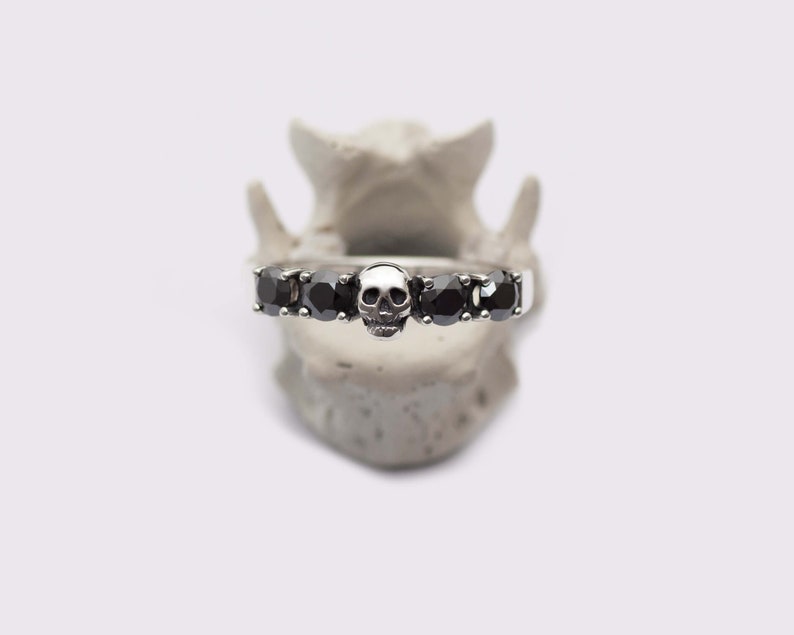May include: A silver band ring with five black gemstones and a skull design in the center.
