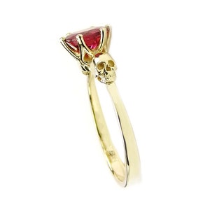 May include: A gold ring with a red gemstone and a skull design. The skull is on the band of the ring, below the gemstone.