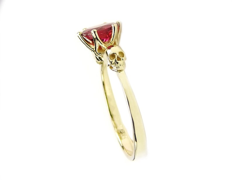 May include: A gold ring with a red gemstone and a skull design. The skull is on the band of the ring, below the gemstone.