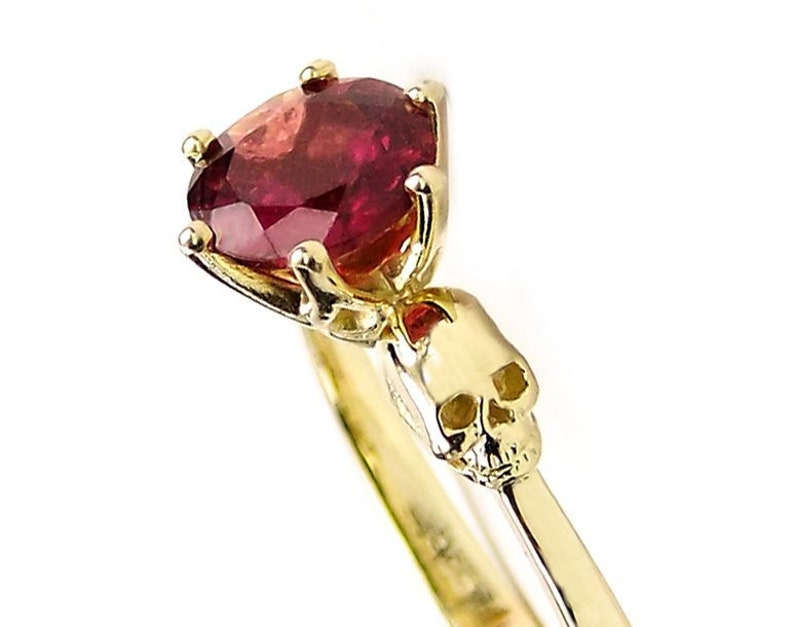 May include: A gold ring with a red gemstone and a skull design. The ring is set with a round, red gemstone in a prong setting. The skull is on the band of the ring and is facing forward.