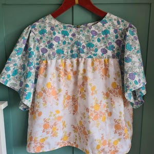 Könnte beinhalten: Eine kurzärmlige Bluse mit floralem Muster. Der obere Teil zeigt einen blauen, lila und weißen Blumendruck, während der untere Teil einen weißen Hintergrund mit orangefarbenen und gelben Blumen hat. Die Bluse hängt an einem Holzbügel.