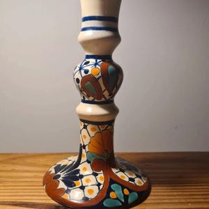 Könnte beinhalten: Ein dekorativer Kerzenhalter aus Keramik mit breitem Sockel und hohem, schlankem Körper. Der Kerzenhalter hat einen cremefarbenen Hintergrund mit aufwendigen Blumenmustern in Orange, Blau und Grün. Die Oberseite ist dunkelblau mit cremefarbenen Streifen.