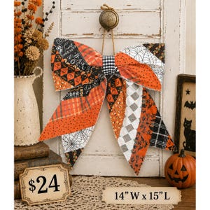 May include: A decorative fabric bow featuring a patchwork design of orange, black, and white Halloween-themed patterns, including spiderwebs, pumpkins, and candy corn. The bow measures 14 inches wide by 15 inches long and hangs from a jute twine loop.