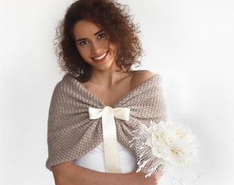 May include: A beige knitted shawl with a cream-colored bow and ribbon detail. The shawl is draped over the shoulders. A white bouquet with a veil is held in the hand.