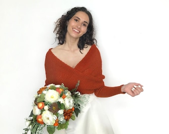 May include: A woman wearing a rust-colored knitted sweater with a white bridal gown and holding a bouquet of white and orange flowers.