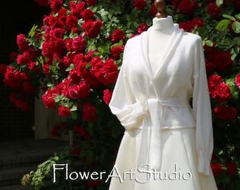 Könnte beinhalten: Ein weißer Strickcardigan mit Schalkragen und einem Bindegürtel. Der Cardigan wird auf einer Schaufensterpuppe vor einem Hintergrund aus roten Rosen präsentiert.