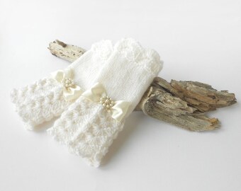 May include: A pair of white fingerless gloves featuring a textured crochet pattern on the lower section and a smooth knit upper section. Each glove is adorned with a cream-colored satin ribbon bow centered with a cluster of small white faux pearls.