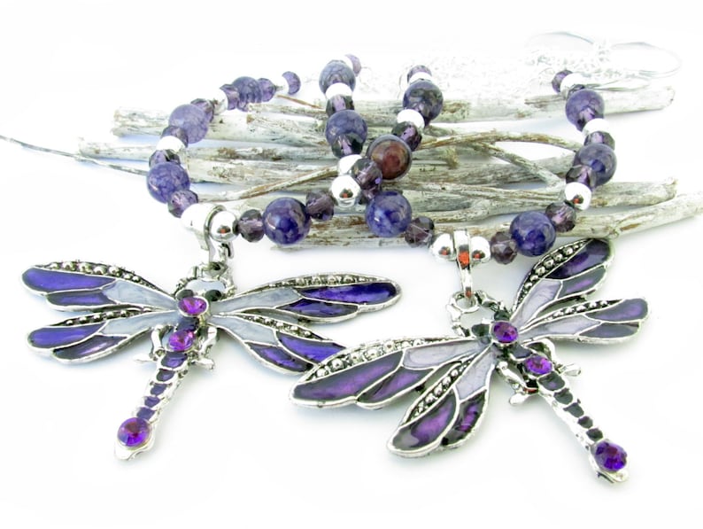 May include: Two dragonfly pendant necklaces. Each necklace features a silver-toned dragonfly pendant with purple and gray enamel wings and purple gemstone accents. The beaded necklace includes purple and silver beads.