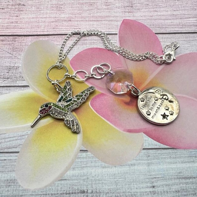 May include: A silver charm necklace with a hummingbird charm, a crystal charm, and a round charm with the text "I love you to the moon & back".