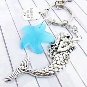 May include: A silver-toned mermaid keychain with a light blue starfish charm and a tag that reads "Dreaming of the Sea." The mermaid has detailed scales and flowing hair. The keychain is attached to a silver ring.