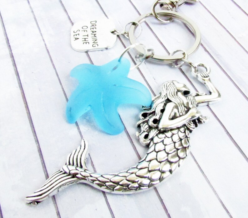 May include: A silver-toned mermaid keychain with a light blue starfish charm and a tag that reads "Dreaming of the Sea." The mermaid has detailed scales and flowing hair. The keychain is attached to a silver ring.