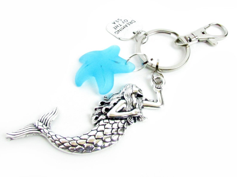 May include: A silver-toned mermaid keychain with a blue starfish charm and a silver clasp. The mermaid has detailed scales and flowing hair. A small tag reads "Dreaming of the Sea".