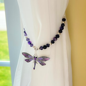 May include: A decorative curtain tieback featuring a purple dragonfly pendant. The dragonfly has detailed wings and is adorned with rhinestones. The tieback is made of a beaded chain with purple and silver beads, adding a touch of elegance to any window treatment.