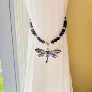 May include: A decorative curtain tieback featuring a dragonfly pendant. The dragonfly has purple and pink wings and a silver-colored body. The tieback is made of purple beads and hangs from a white curtain.