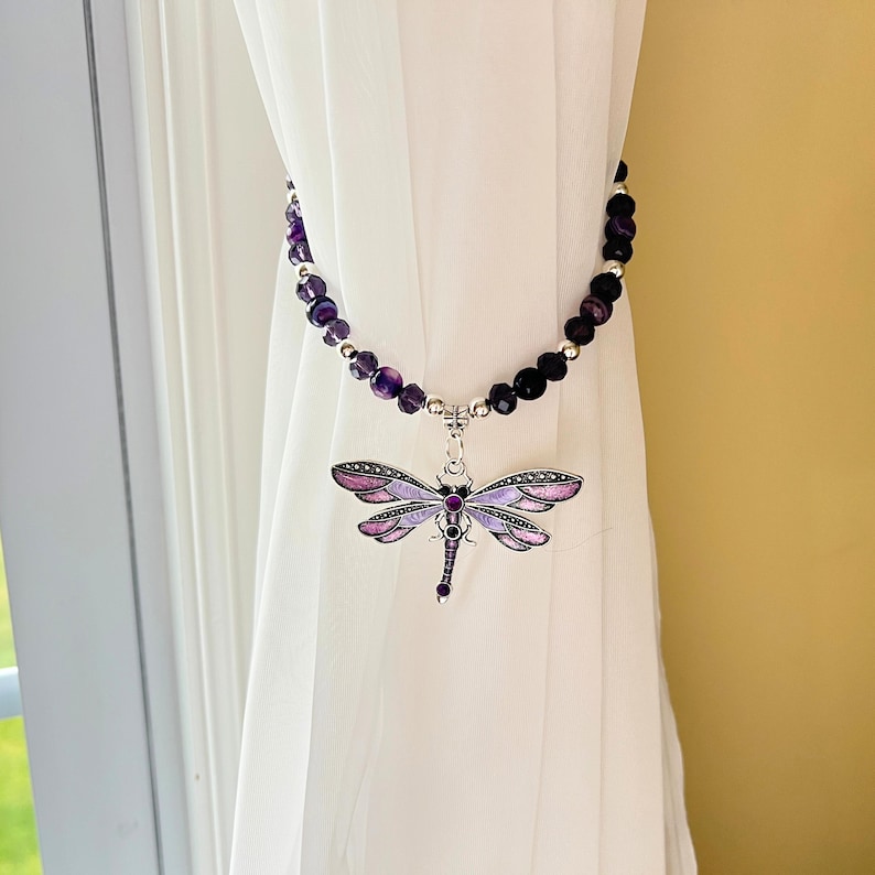 May include: A decorative curtain tieback featuring a dragonfly pendant. The dragonfly has purple and pink wings and a silver-colored body. The tieback is made of purple beads and hangs from a white curtain.