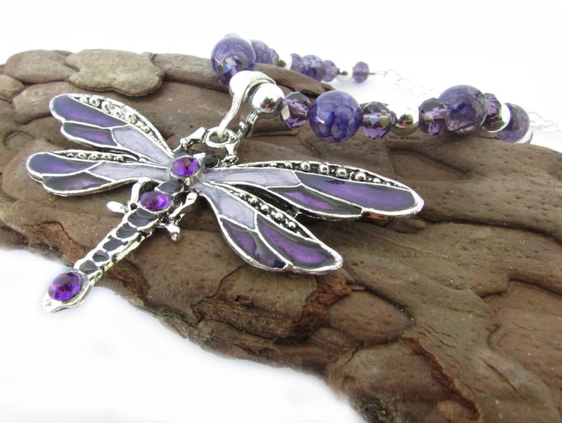 May include: A silver-toned dragonfly pendant necklace with purple enamel wings and purple gemstone accents. The necklace features a beaded strand of purple and silver beads. The dragonfly pendant is approximately 3 inches long.