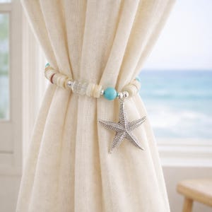 May include: A curtain tieback featuring a strand of off-white disc-shaped beads, clear glass beads, and bright turquoise round beads. A silver-toned metal starfish charm hangs from the center of the beaded strand, which is wrapped around a curtain.