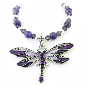 May include: A silver-toned dragonfly pendant necklace with purple and lavender enamel wings and a matching beaded chain. The dragonfly has a detailed design with small crystals and purple gemstone accents. The necklace is a jewelry item.