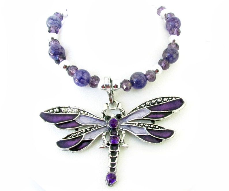 May include: A silver-toned dragonfly pendant necklace with purple and lavender enamel wings and a matching beaded chain. The dragonfly has a detailed design with small crystals and purple gemstone accents. The necklace is a jewelry item.