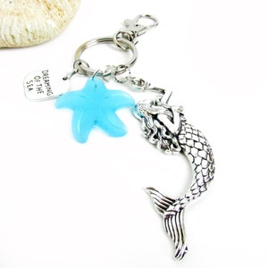 May include: A silver-toned keychain featuring a mermaid charm, a light blue starfish, and a tag that reads "Dreaming of the Sea." The mermaid charm has detailed scales and flowing hair. The keychain includes a clasp and a key ring.