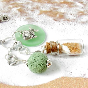 May include: A green glass charm with a silver fish charm, a green coral bead, and a small glass bottle filled with sand and a seashell. The charms are attached to a silver chain.