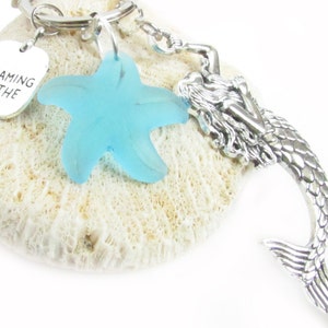 May include: A silver keychain featuring a mermaid charm, a light blue starfish, and a small tag that reads "Dreaming of the Sea." The mermaid charm is detailed with scales and flowing hair. The starfish is a translucent blue.