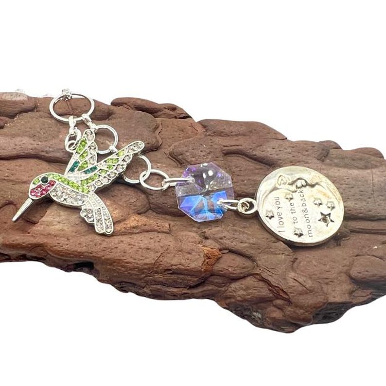 May include: A silver charm bracelet with a hummingbird charm, a crystal charm, and a moon charm with the text "I love you to the moon & back".