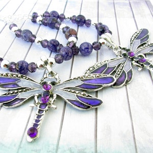 May include: A silver necklace with a dragonfly pendant. The pendant is purple, gray, and silver with purple gemstone accents. The necklace also features purple and silver beads.
