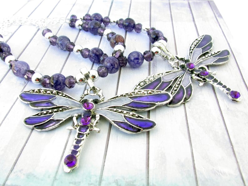 May include: A silver necklace with a dragonfly pendant. The pendant is purple, gray, and silver with purple gemstone accents. The necklace also features purple and silver beads.