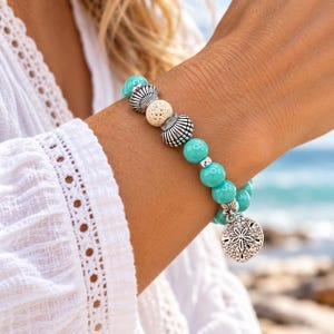 May include: A stretch bracelet featuring smooth turquoise-colored beads, silver-toned metal shell-shaped spacers, a single porous beige lava stone bead, and a round silver-toned metal charm embossed with a detailed sand dollar design.