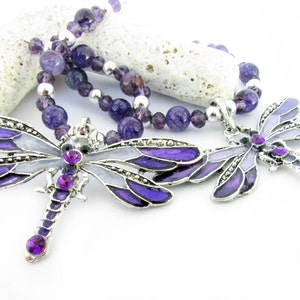 May include: A necklace featuring two dragonfly pendants with purple and silver accents. The dragonfly pendants have purple enamel wings and are adorned with purple gemstones. The necklace also includes a beaded strand of purple and silver beads.