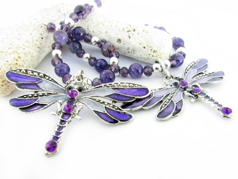 May include: A necklace featuring two dragonfly pendants with purple and silver accents. The dragonfly pendants have purple enamel wings and are adorned with purple gemstones. The necklace also includes a beaded strand of purple and silver beads.