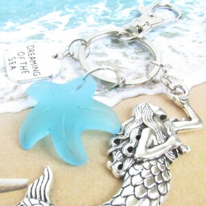 May include: A silver keychain with a mermaid charm, a light blue starfish, and a tag that reads "Dreaming of the Sea." The keychain is on a sandy beach background with ocean waves.