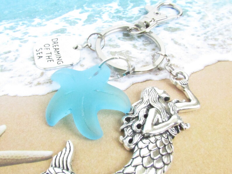 May include: A silver keychain with a mermaid charm, a light blue starfish, and a tag that reads "Dreaming of the Sea." The keychain is on a sandy beach background with ocean waves.