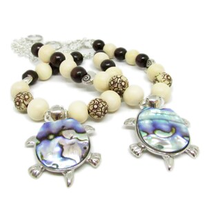 May include: Two beaded necklaces with a silver turtle pendant. The beads are brown, white, and cream colored. The turtle pendant has a iridescent abalone shell.