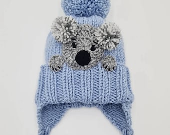 May include: A light blue knitted hat with a koala face and a fluffy pom pom on top. The hat has ear flaps that tie under the chin.