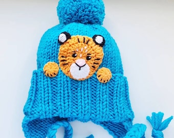 May include: Blue knitted hat with a tiger face applique and a fluffy pom pom on top. The hat has ear flaps that tie under the chin.