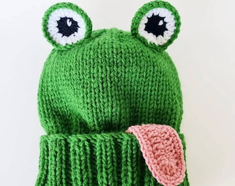 May include: Green knitted frog hat with a pink crocheted tongue. The hat has white and black eyes.