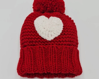 May include: Red knitted beanie with a white heart on the front and a red pom pom on top.