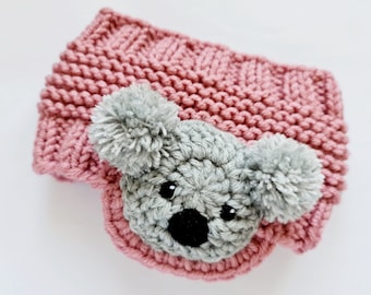 May include: A pink knitted headband with a gray koala face and pom-poms on the ears.