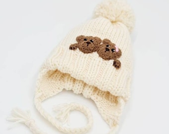 May include: Cream-colored knit hat with ear flaps and a pom-pom. Two brown teddy bear appliqués are on the front. The hat has braided ties with tassels. A small pink flower is on one of the bears.